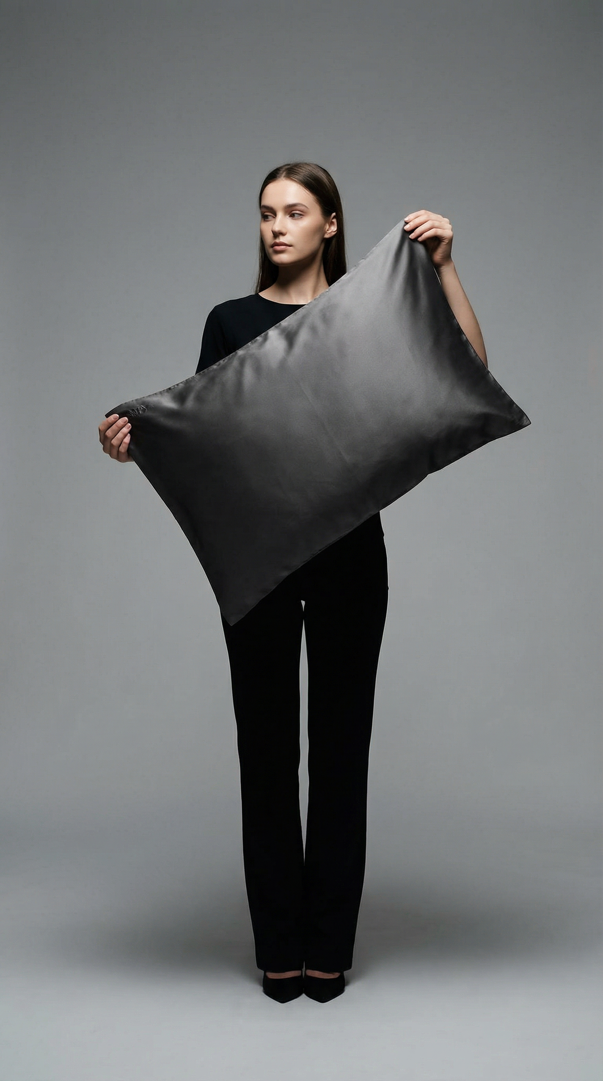 Stylish woman in black holding large gray pillow on plain background